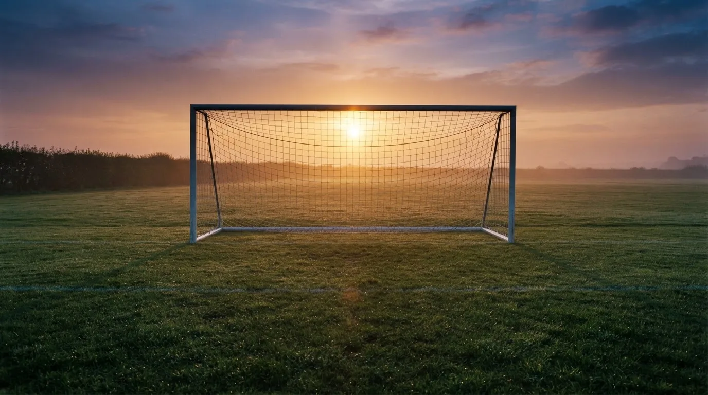 Ein Fußballtor mit Netz bei Sonnenuntergang auf einem leeren Platz, die Schatten der Torpfosten liegen lang auf dem Gras.