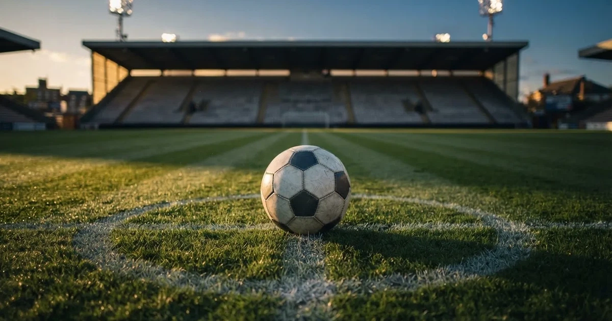 Ein Fußball liegt auf dem Anstoßpunkt eines leeren Rasenplatzes, im Hintergrund eine große Stadiontribüne im Abendlicht.