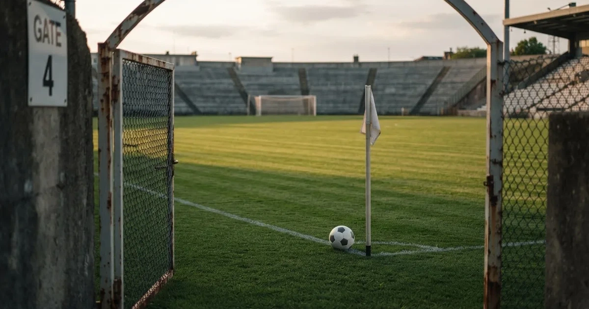 Blick durch ein Stadiontor auf einen grünen Rasen im Abendlicht, ein Ball ruht an der Eckfahne, die Tribünen sind leer.