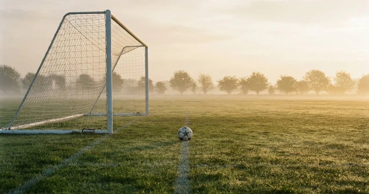 Ein Fußballtor mit gespanntem Netz steht auf einem leeren Rasenplatz, davor ein einzelner Ball, Morgendunst liegt über dem Feld.