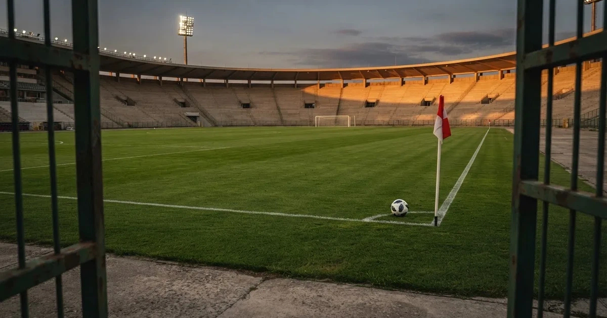 Blick durch ein Stadiontor auf einen grünen Rasen im Abendlicht, ein Ball liegt an der Eckfahne, die Ränge sind leer.