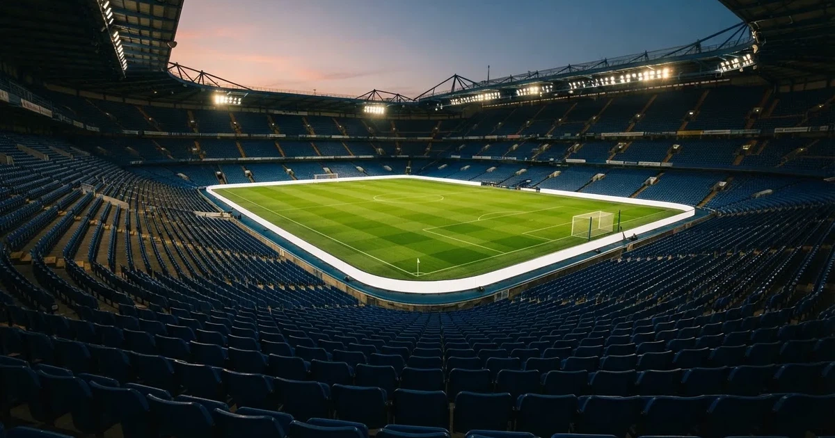 Leere Sitzreihen eines großen Stadions unter Flutlicht, dahinter die ferne Sicht auf den hell beleuchteten Rasen nach Sonnenuntergang.
