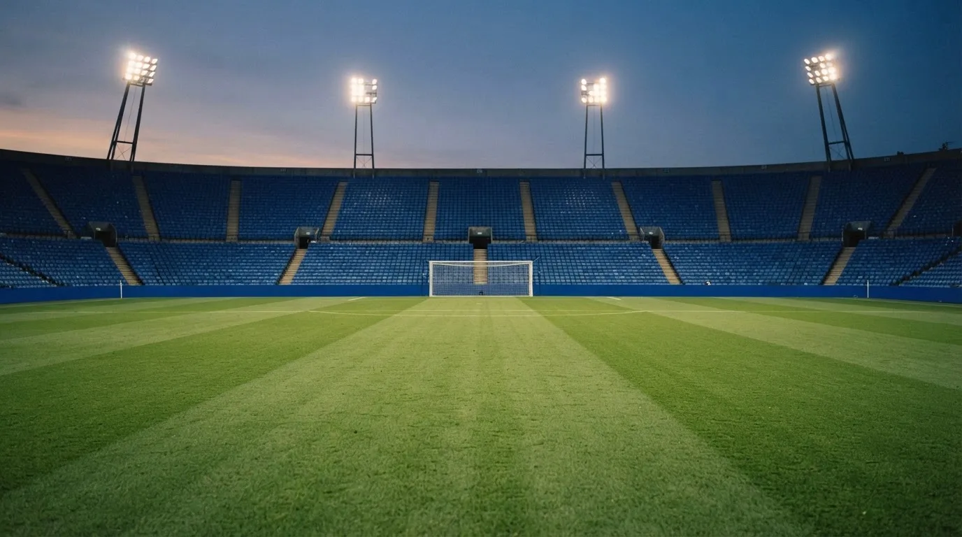 Eine leere Stadiontribüne unter Flutlicht aus der Ferne betrachtet, die Rasenfläche leuchtet grün in der Dämmerung.