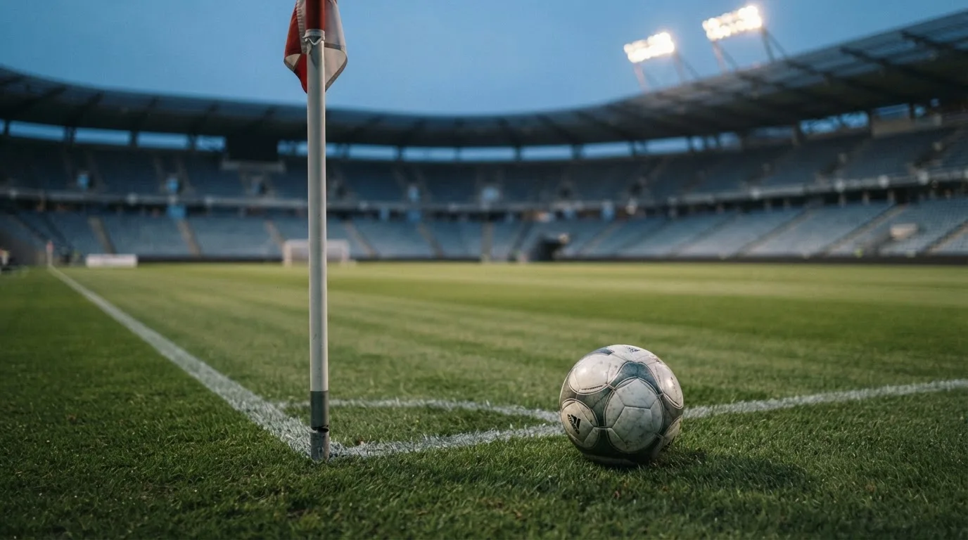 Ein Fußball liegt neben einem Eckfahnen-Pfosten auf einem Rasenplatz, im Hintergrund ein leeres Stadion in der Dämmerung.