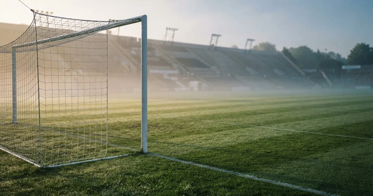 Ein Fußballplatz mit gepflegtem Rasen und einem Tor, über dem Spielfeld liegt Morgennebel, im Hintergrund eine leere Tribüne.