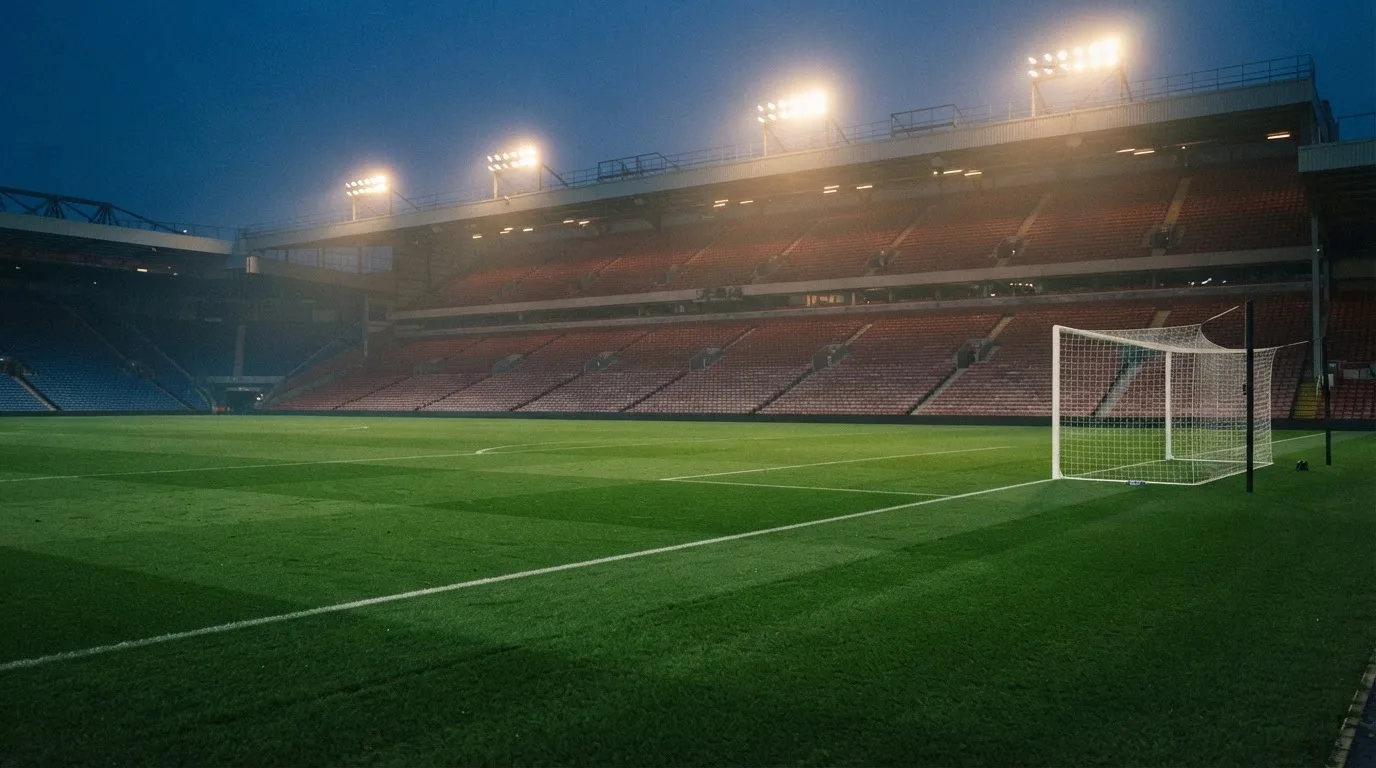 Eine leere Stadiontribüne unter Flutlicht aus der Ferne betrachtet, die Rasenfläche leuchtet grün in der Dämmerung.