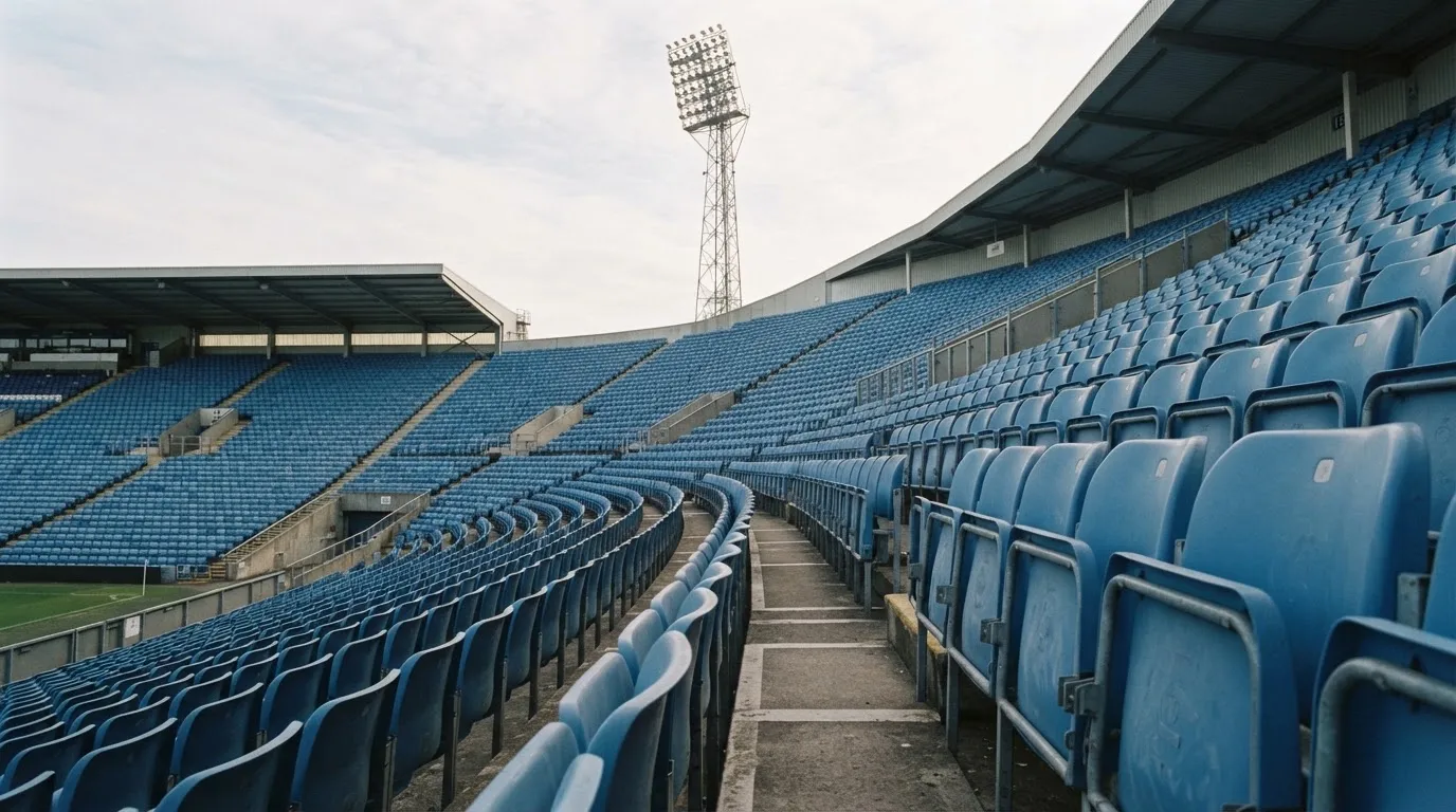 Eine leere Tribüne eines Fußballstadions bei Tageslicht, Sitzschalen in Reihen und dahinter ein Flutlichtmast.