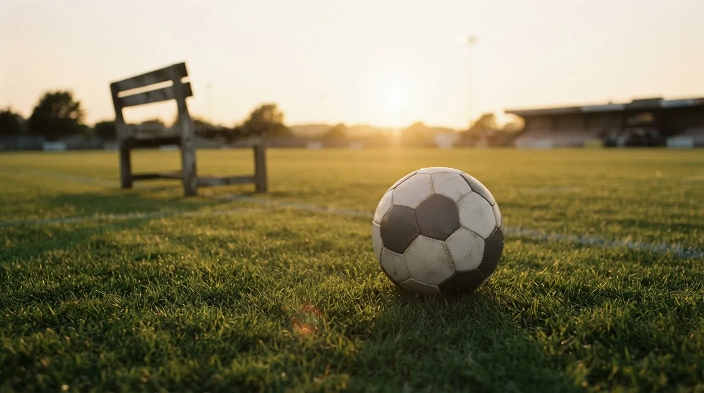 Ein Fußball auf einem Rasenplatz im Gegenlicht der tiefstehenden Sonne, am Rand eine leere Ersatzbank.
