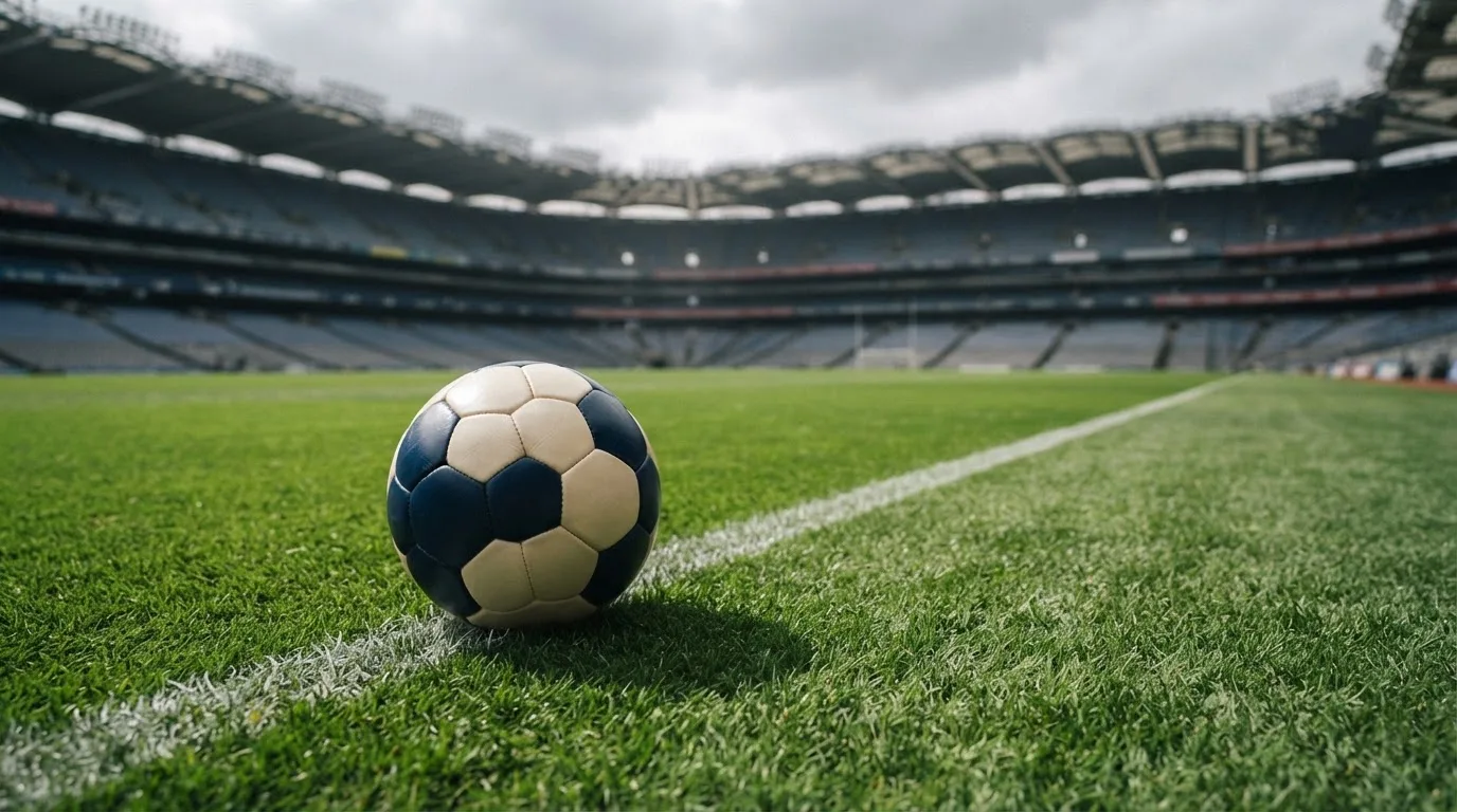 Ein Fußball auf einem Rasen, im Hintergrund ein leeres Stadion bei bewölktem Himmel.