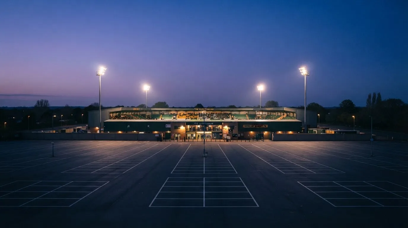 Ein Fußballstadion unter Flutlicht, aus großer Entfernung bei Einbruch der Dunkelheit gesehen, davor ein leerer Parkplatz.