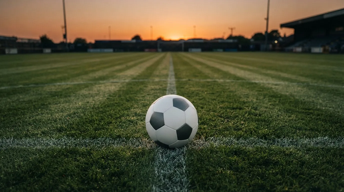 Ein Fußball liegt in der Mitte eines leeren Rasenplatzes, am Horizont färbt sich der Himmel orange.
