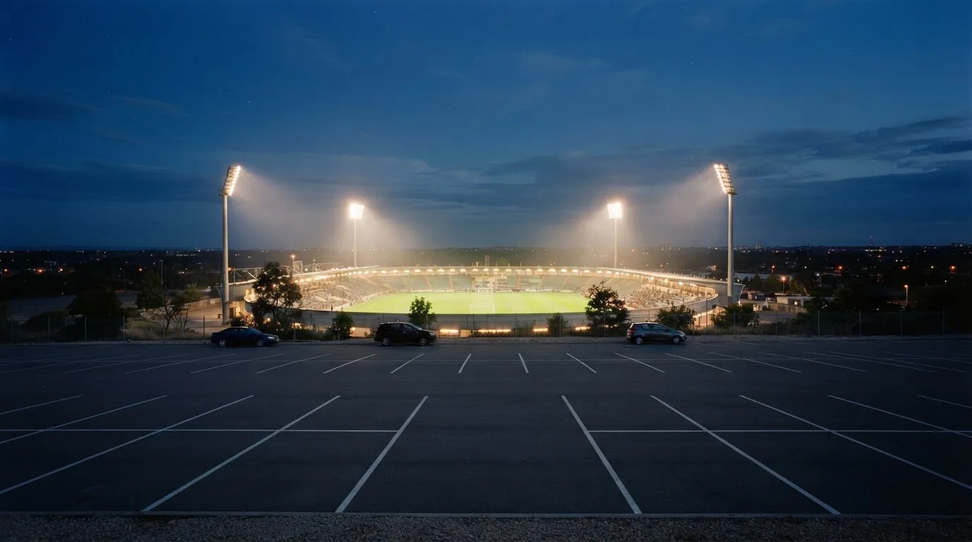 Ein Stadion unter Flutlicht aus großer Entfernung bei Einbruch der Dunkelheit, davor ein stiller Parkplatz.