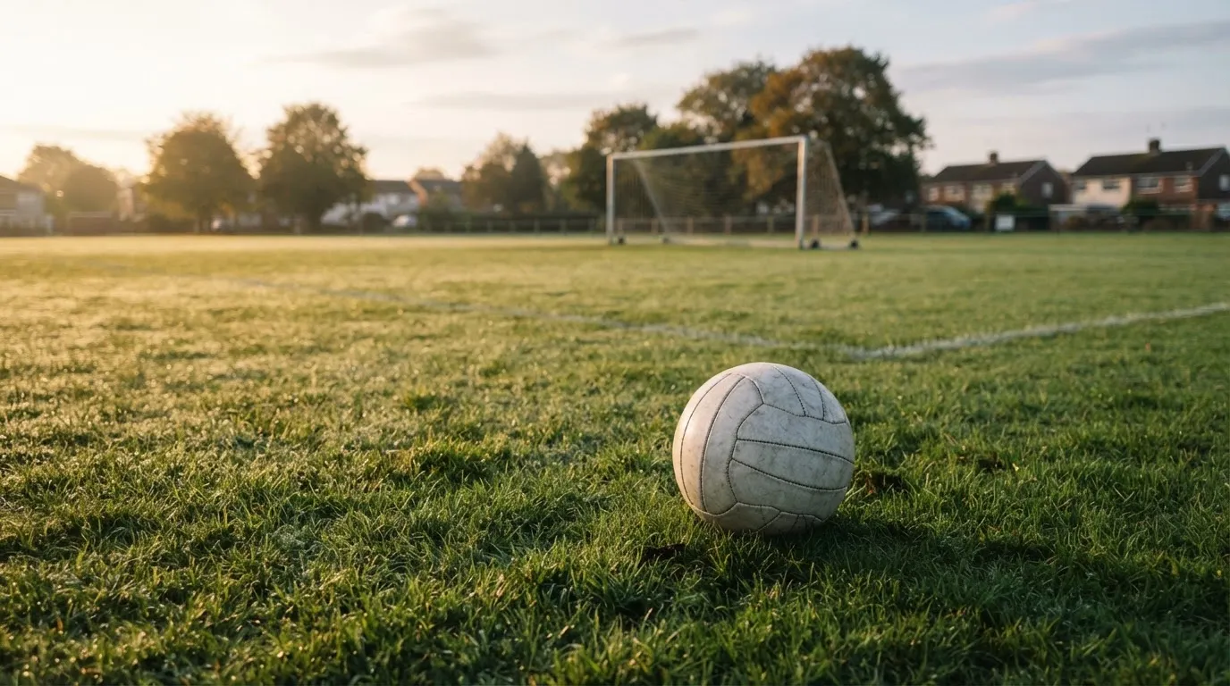 Ein Fußball auf dem Rasen eines leeren Sportplatzes am Rand einer Kleinstadt im Morgenlicht.