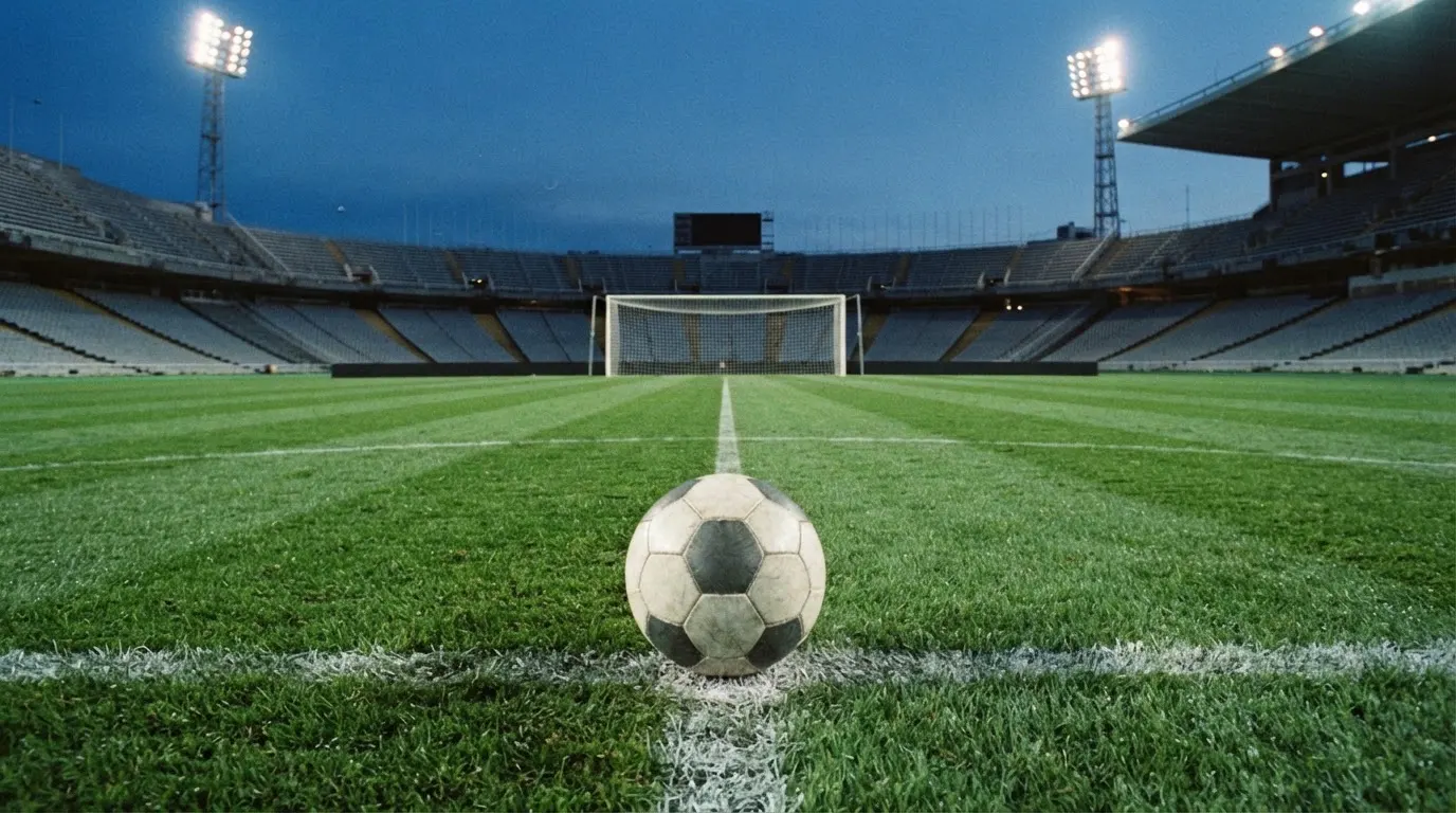 Ein Fußball liegt auf dem Anstoßpunkt eines leeren Rasenplatzes, im Hintergrund ein Stadion unter Flutlicht in der Abenddämmerung.