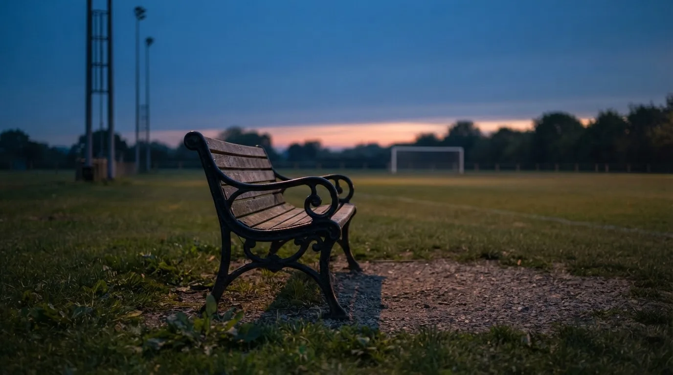 Eine leere Parkbank am Rand eines Fußballfelds in der Abenddämmerung, im Hintergrund ragen dunkle Flutlichtmasten in den Himmel.