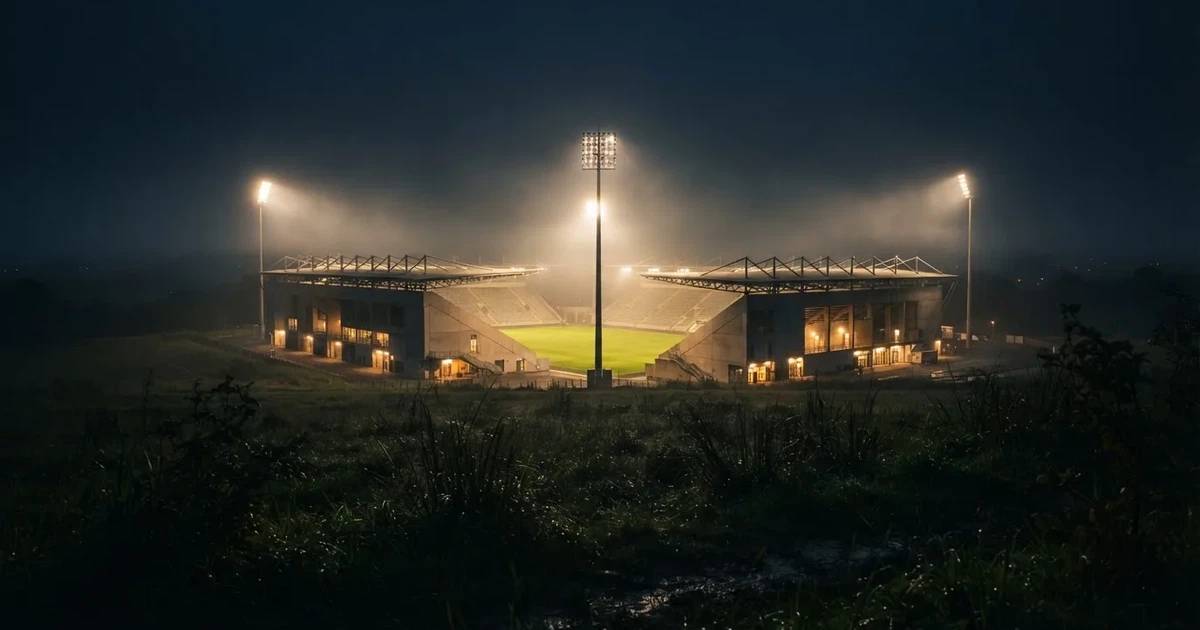 Ein Stadion unter Flutlicht, aus großer Entfernung über eine dunkle Wiese fotografiert, am Himmel zieht Nebel auf.