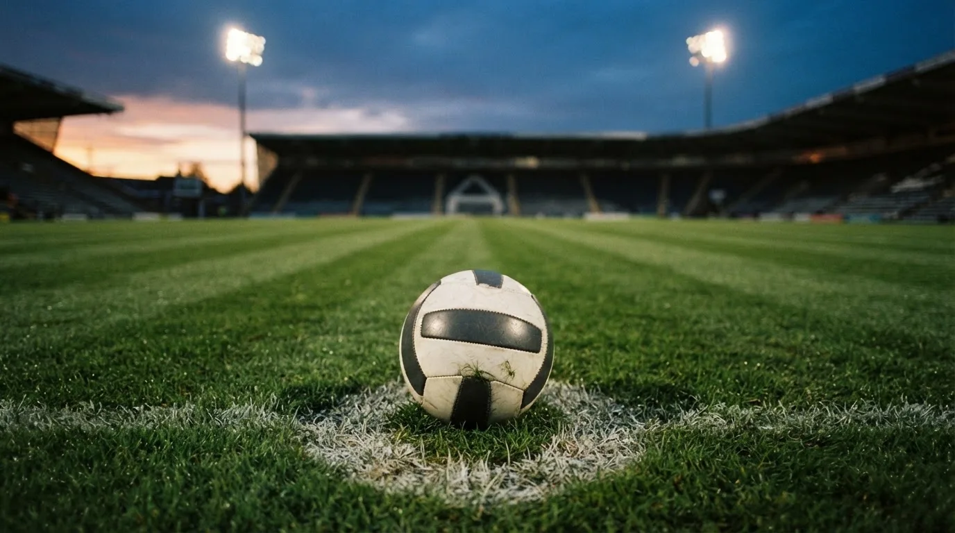 Ein Fußball liegt auf dem Anstoßpunkt eines leeren Rasenplatzes, im Hintergrund steht ein Stadion unter Flutlicht in der Dämmerung.