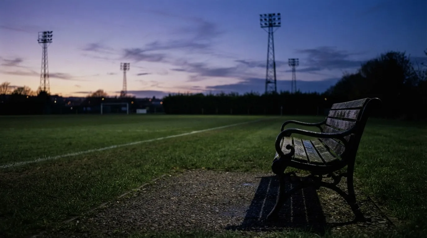 Eine leere Parkbank am Rand eines Fußballfelds in der Abenddämmerung, im Hintergrund ragen dunkle Flutlichtmasten in den Himmel.