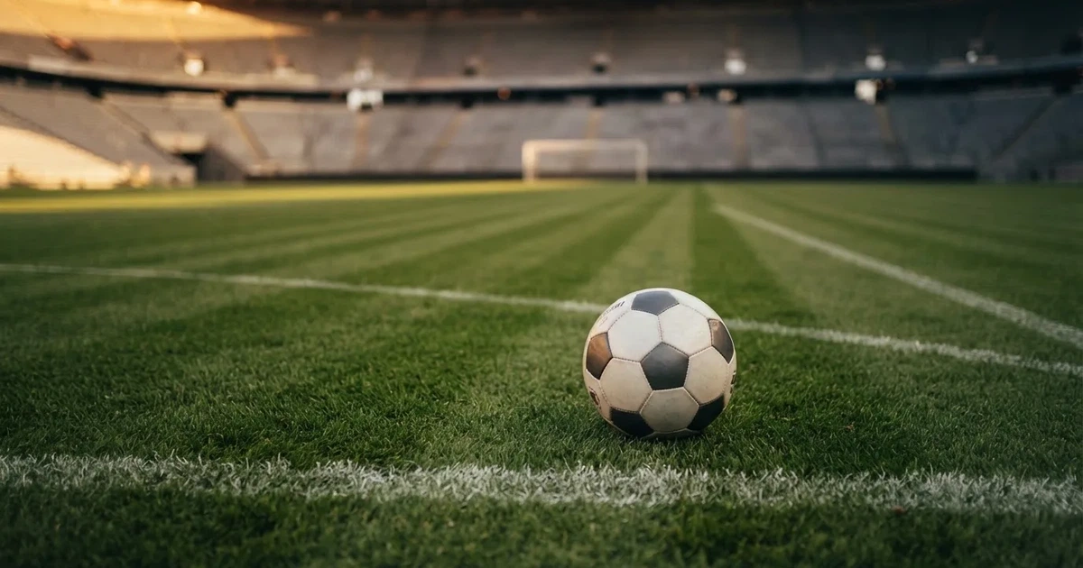 Ein Fußballplatz mit frisch gemähtem Rasen im Abendlicht, ein Ball liegt im Strafraum, im Hintergrund unscharf eine leere Tribüne.