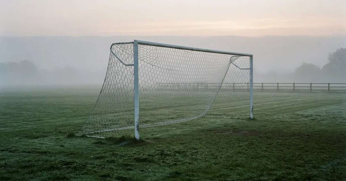 Ein Fußballtor mit Netz steht auf einem leeren Platz im Nebel der Morgendämmerung, der Rasen ist taufeucht.