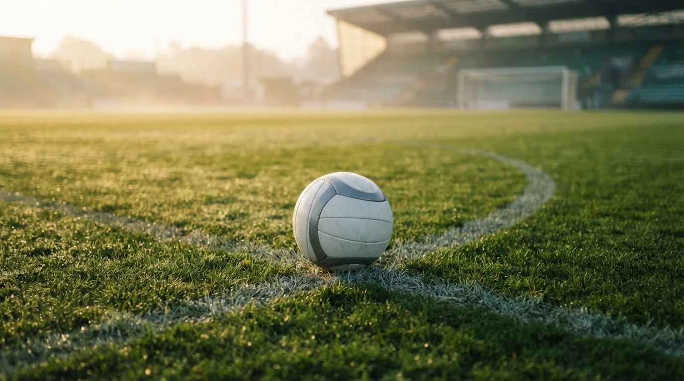 Ein Fußball ruht auf dem Mittelkreis eines leeren Rasenplatzes in der Morgendämmerung, Tau liegt auf dem Gras.