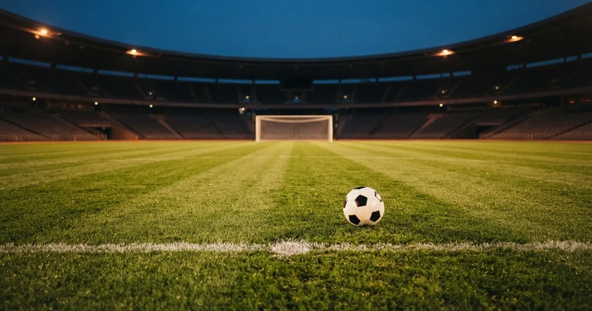 Ein Fußballplatz mit frisch gemähtem Rasen im Abendlicht, ein Ball liegt im Strafraum, im Hintergrund unscharf eine leere Tribüne.