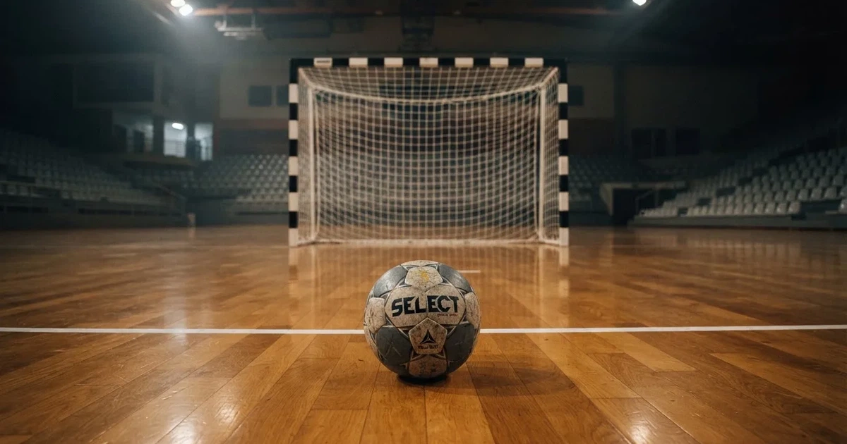 Ein Handball liegt auf dem Hallenboden vor einem Tor mit Netz, die leere Halle wird von Flutlicht beleuchtet.