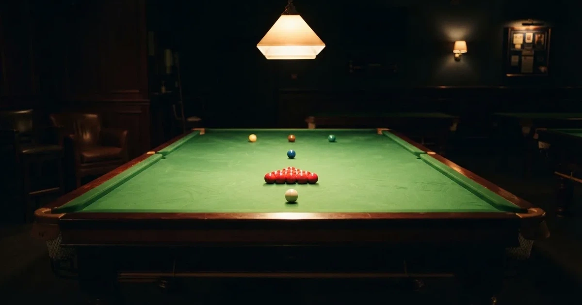 Ein Snookertisch mit grünem Tuch und aufgebauten roten Kugeln in einem abgedunkelten Raum unter einer hängenden Lampe.