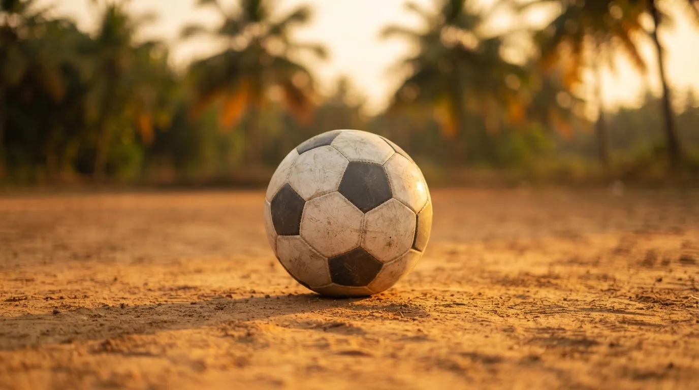 Ein Fußball liegt auf einem staubigen Bolzplatz mit Palmen im Hintergrund in warmem Abendlicht.