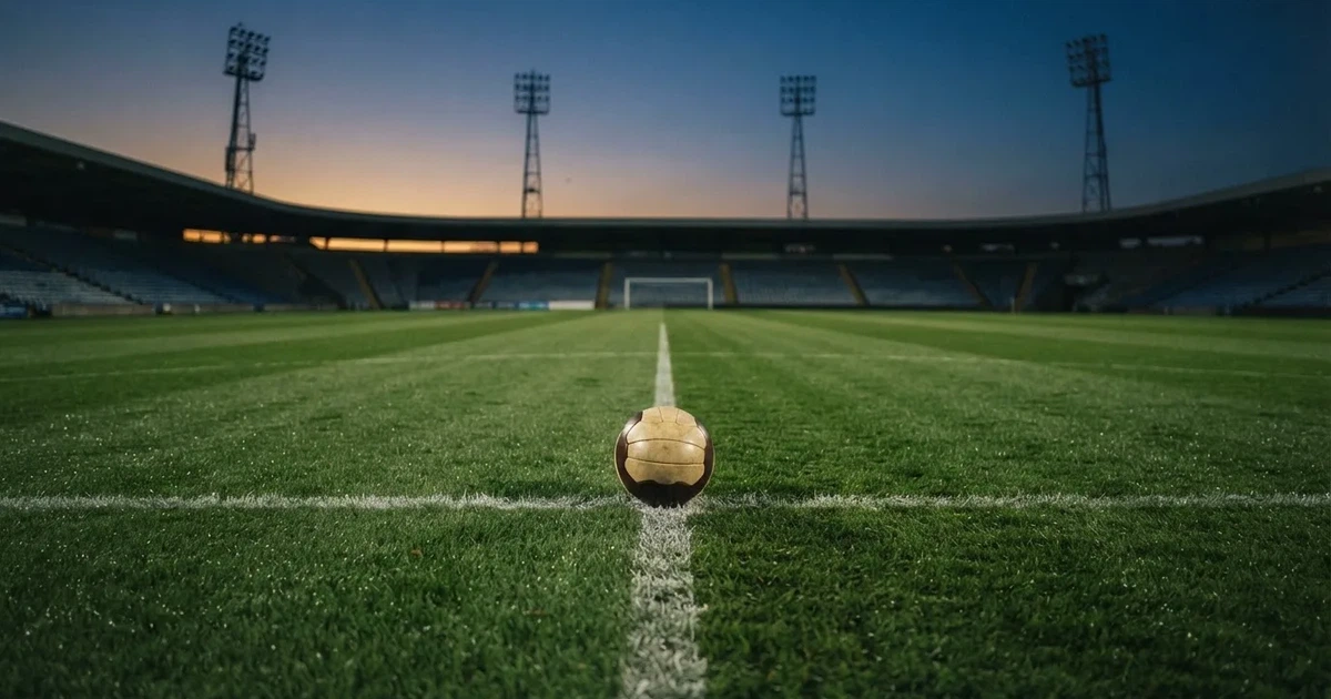 Ein Fußball liegt auf dem Anstoßpunkt eines leeren Rasenplatzes, im Hintergrund eine Tribüne und Flutlichtmasten in der Abenddämmerung.