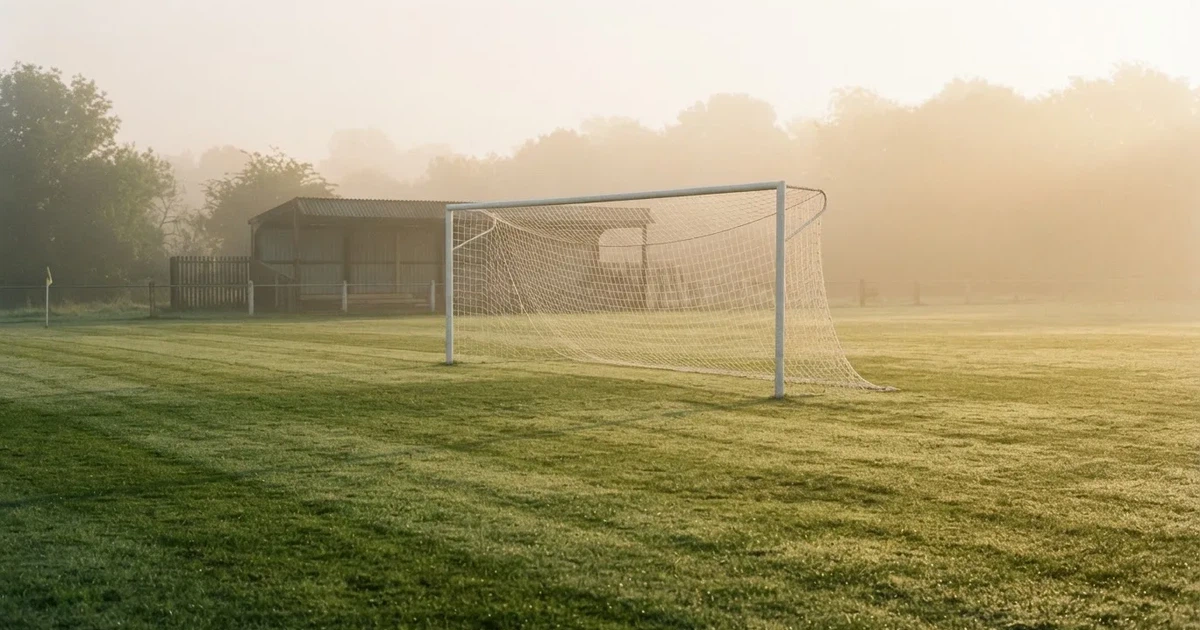 Ein Fußballtor mit Netz steht im Morgennebel auf einem leeren Rasenplatz, im Hintergrund die Silhouette einer kleinen Tribüne.
