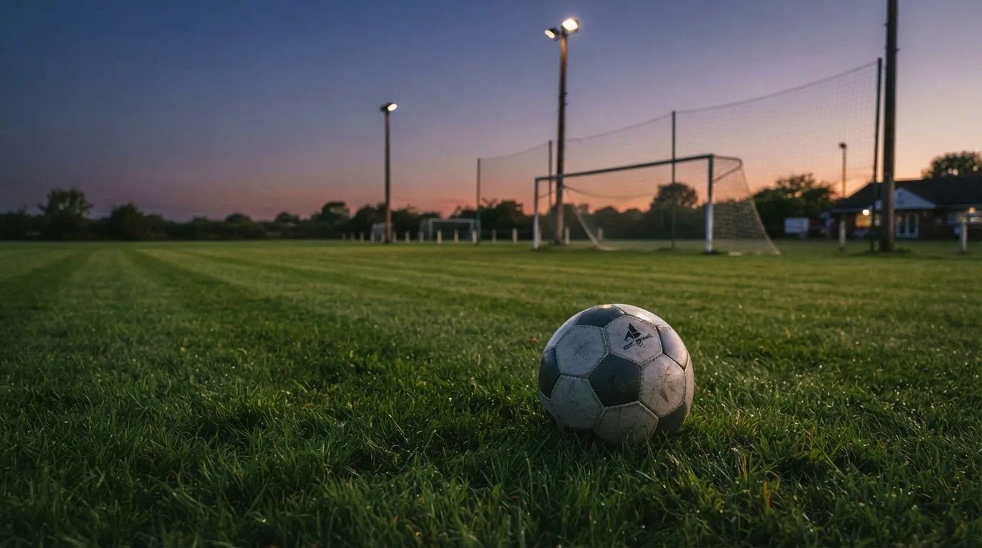 Ein Fußball liegt im Gras eines leeren Platzes, im Hintergrund stehen Flutlichtmasten in der Dämmerung.