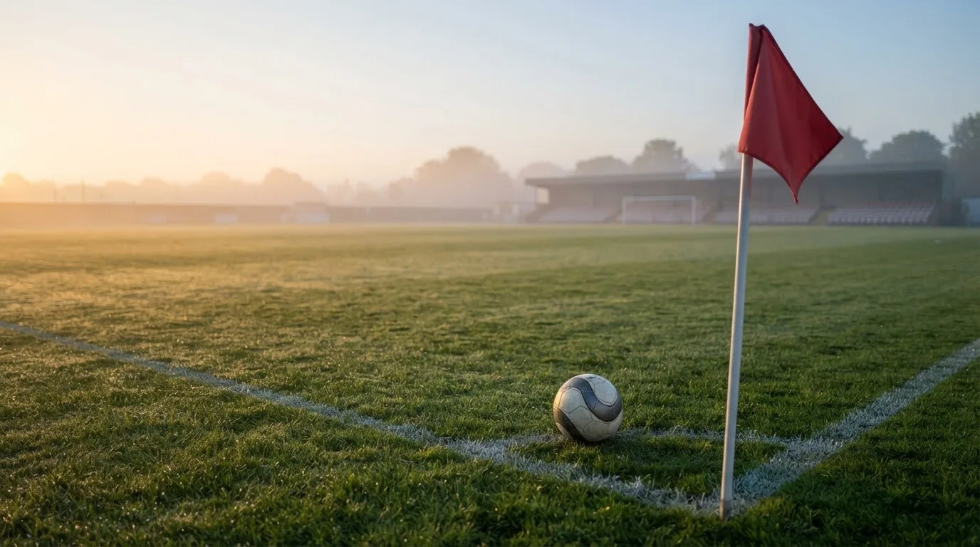 Ein Fußball liegt auf Rasen neben einer Eckfahne ohne Aufdruck auf einem leeren Platz im Morgenlicht.