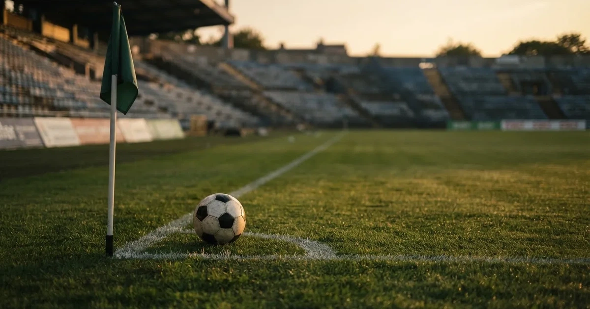 Ein Fußball liegt am Viertelkreis an der Eckfahne eines Rasenplatzes, im Hintergrund leere Tribünen in warmem Abendlicht.