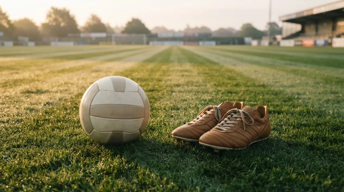 Ein Fußball auf frisch gemähtem Rasen im Morgenlicht, daneben ein Paar Fußballschuhe ohne erkennbare Marke.