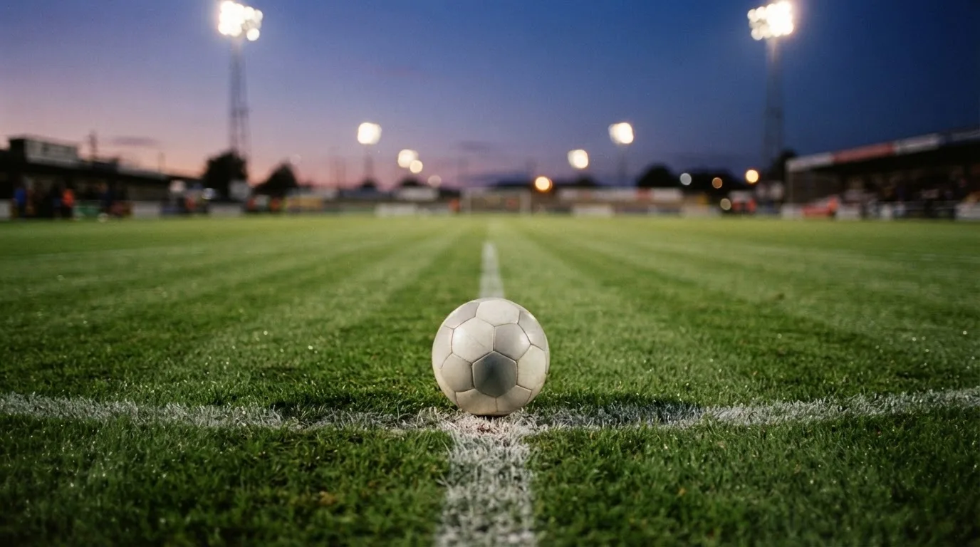 Ein Fußball liegt auf dem Anstoßpunkt eines Rasenplatzes in der Abenddämmerung, Flutlichtmasten stehen unscharf im Hintergrund.