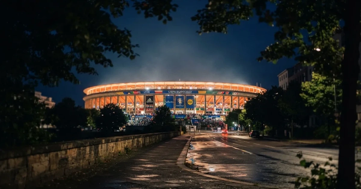 Ein hell erleuchtetes Stadion aus der Ferne bei Nacht, davor eine stille Straße und ein paar Bäume.
