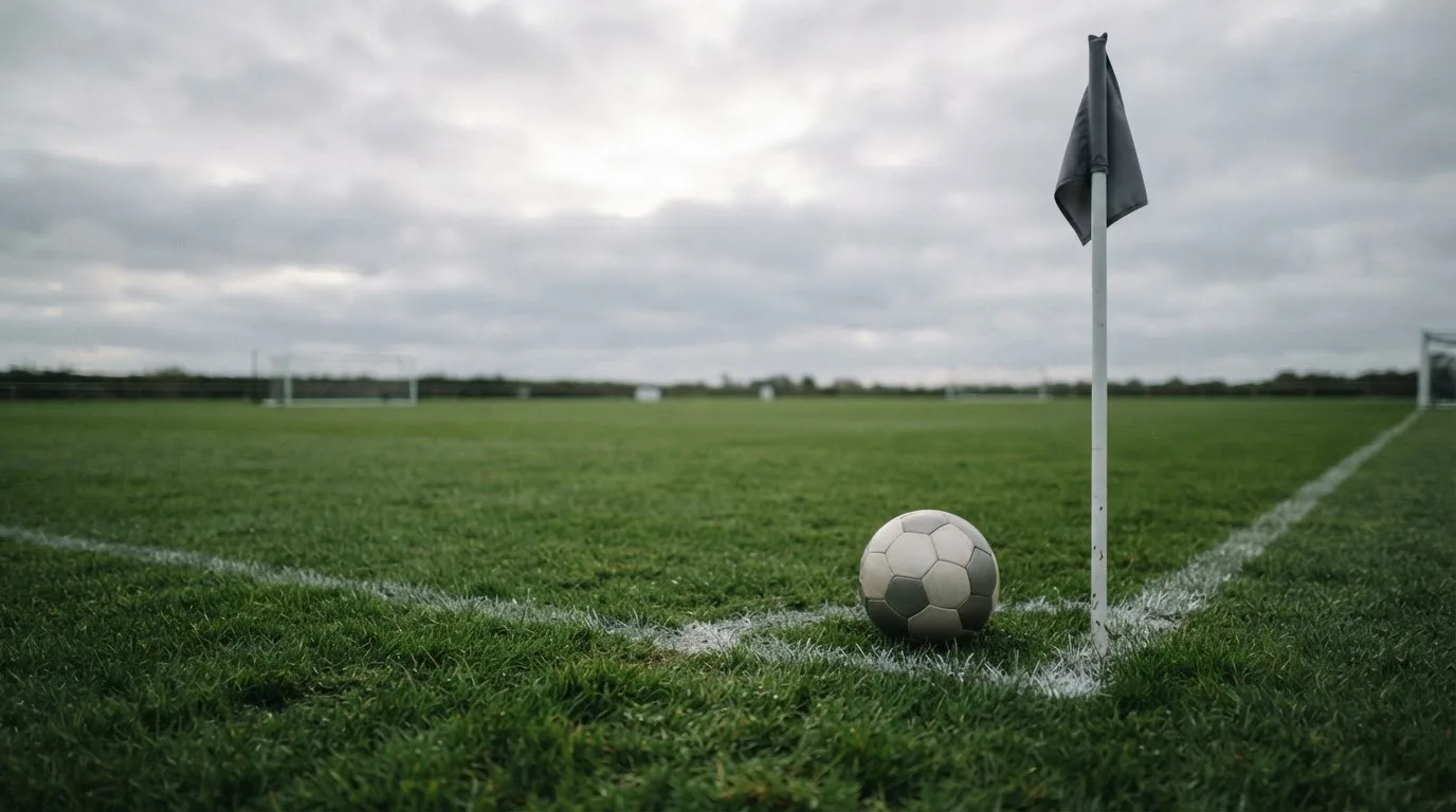 Ein Fußball liegt neben einer Eckfahne auf kurz gemähtem Rasen unter bewölktem Himmel.