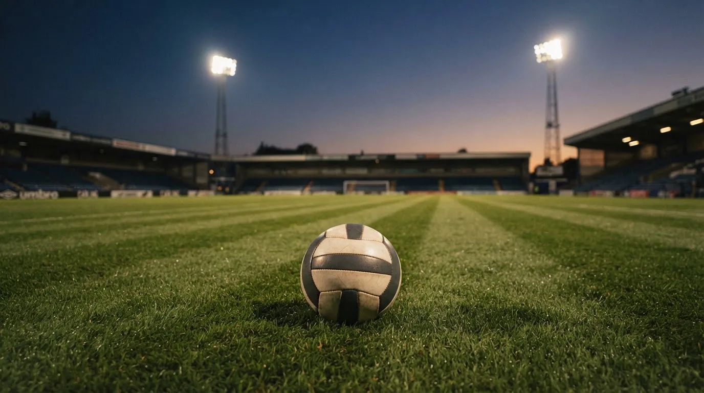 Ein Fußball liegt auf einem Rasenplatz in der Dämmerung, im Hintergrund ein ferner Stadionrand unter Flutlicht.